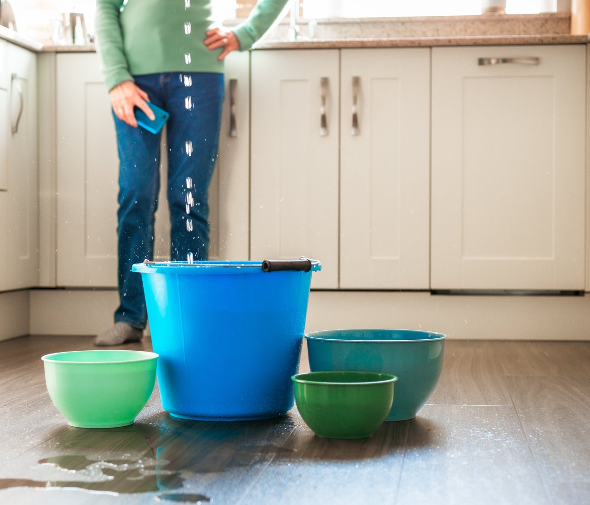 A woman in the background as water leaks in the kitchen ceiling. Buckets and pots are on the floor to catch the flowing water, water puddling on the floor.