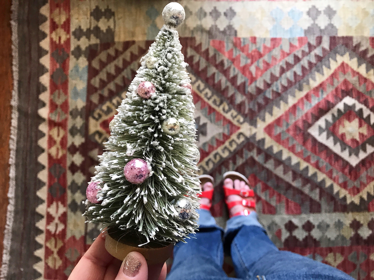 Person holding a small bottle brush Christmas tree in their hand, looking down towards their feet.