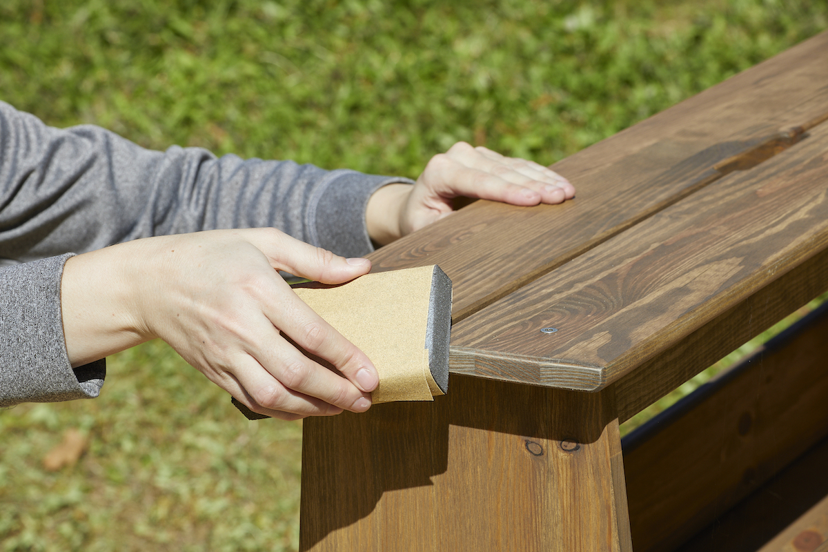 Woman uses a sanding block on a stained wood bench.