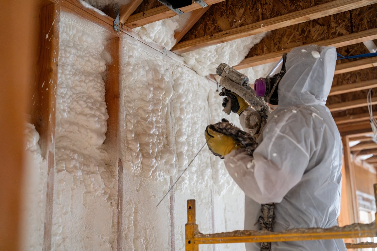 Man wearing protective gear uses a machine to blow insulation into attic walls.