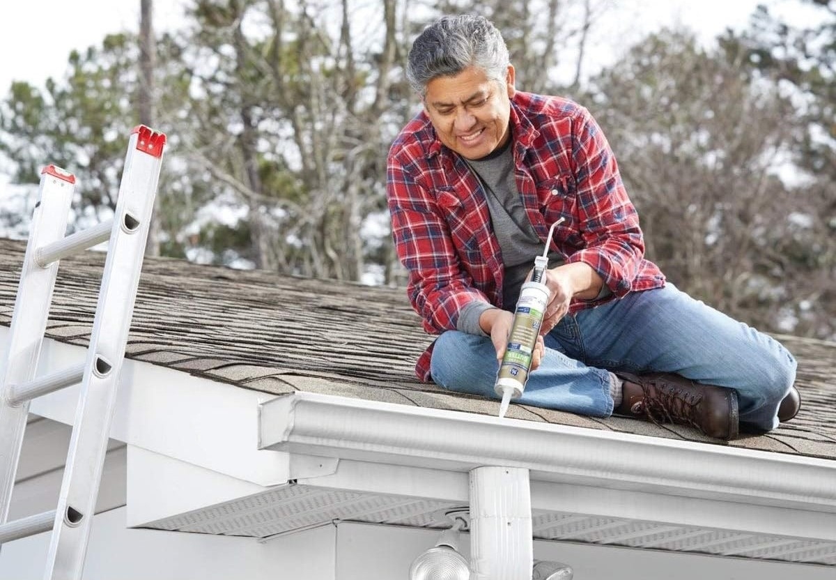 A male homeowner is on the roof using a sealing gun to seal holes in the gutter.