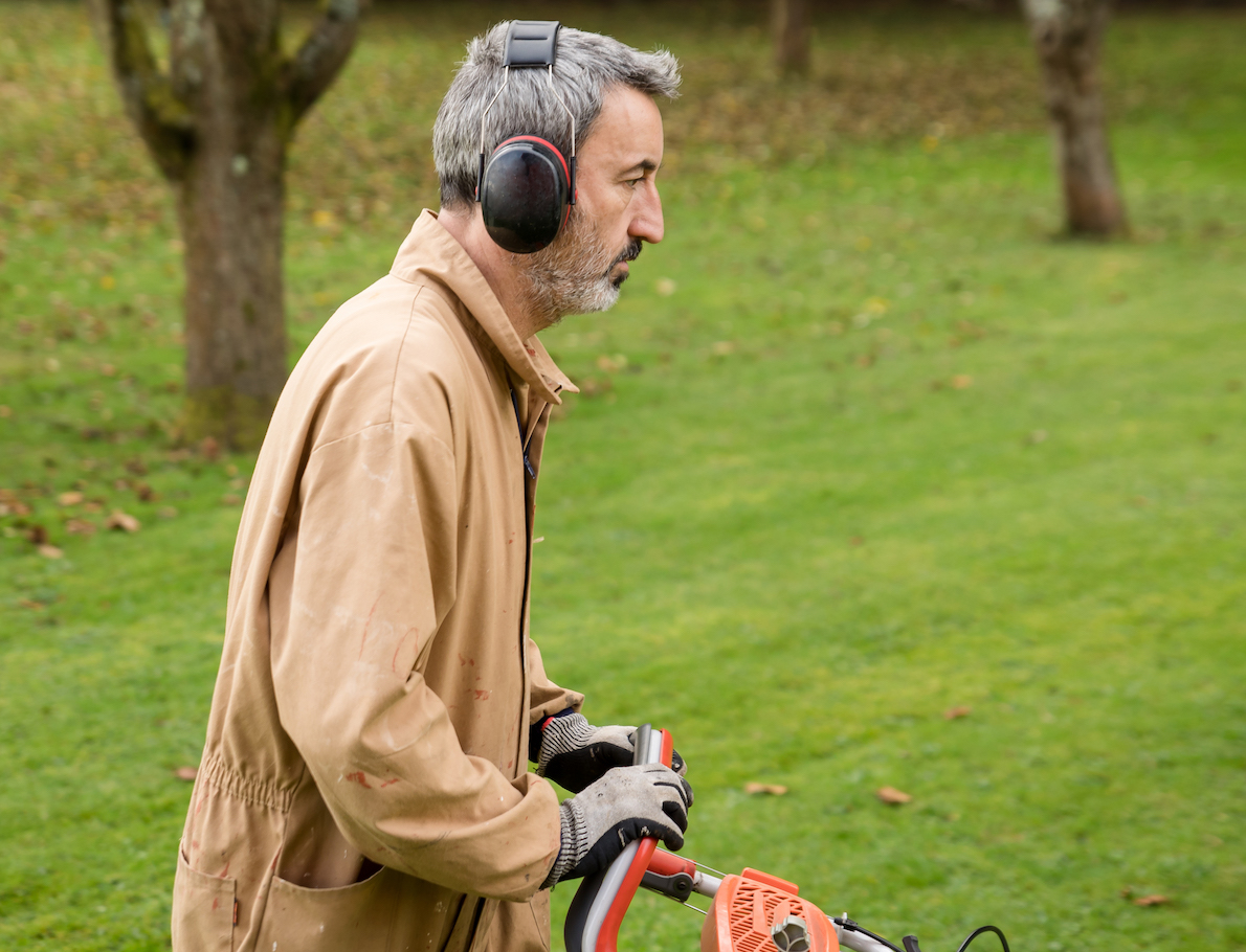 Man in uniform and with ear protectors cutting grass with lawn mower outdoors in a garden. 