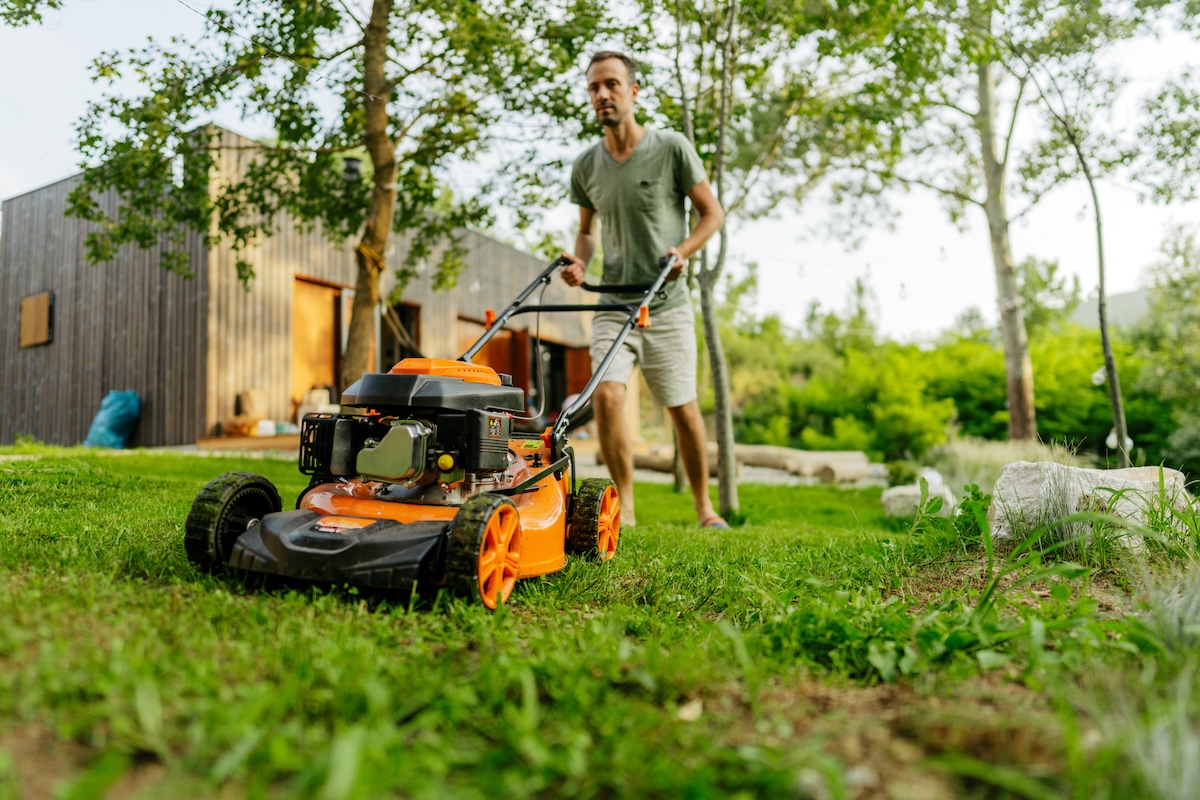 A man in a green shirt pushes an orange gas lawn mower.