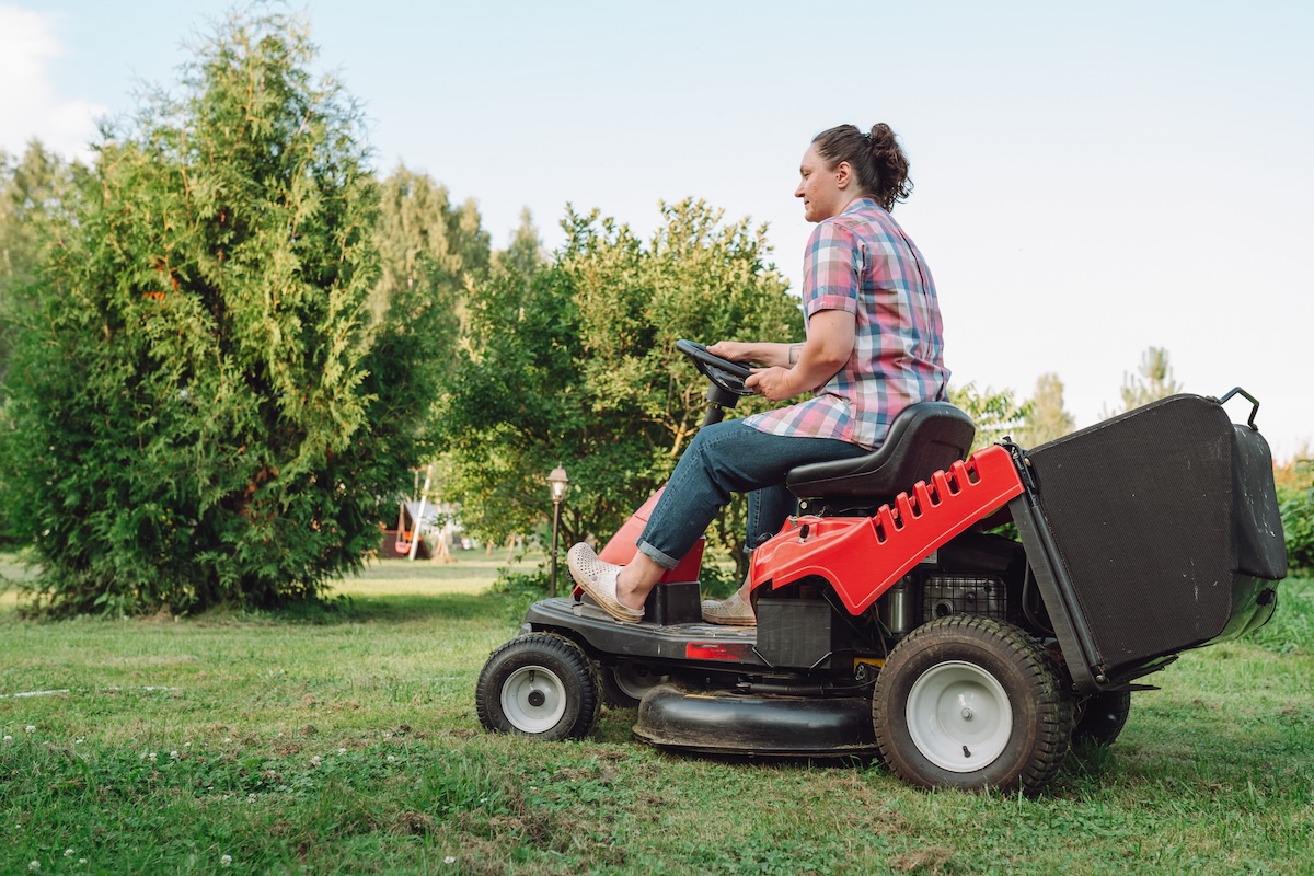 A woman in a plaid shirt rides a red tractor lawn mower.