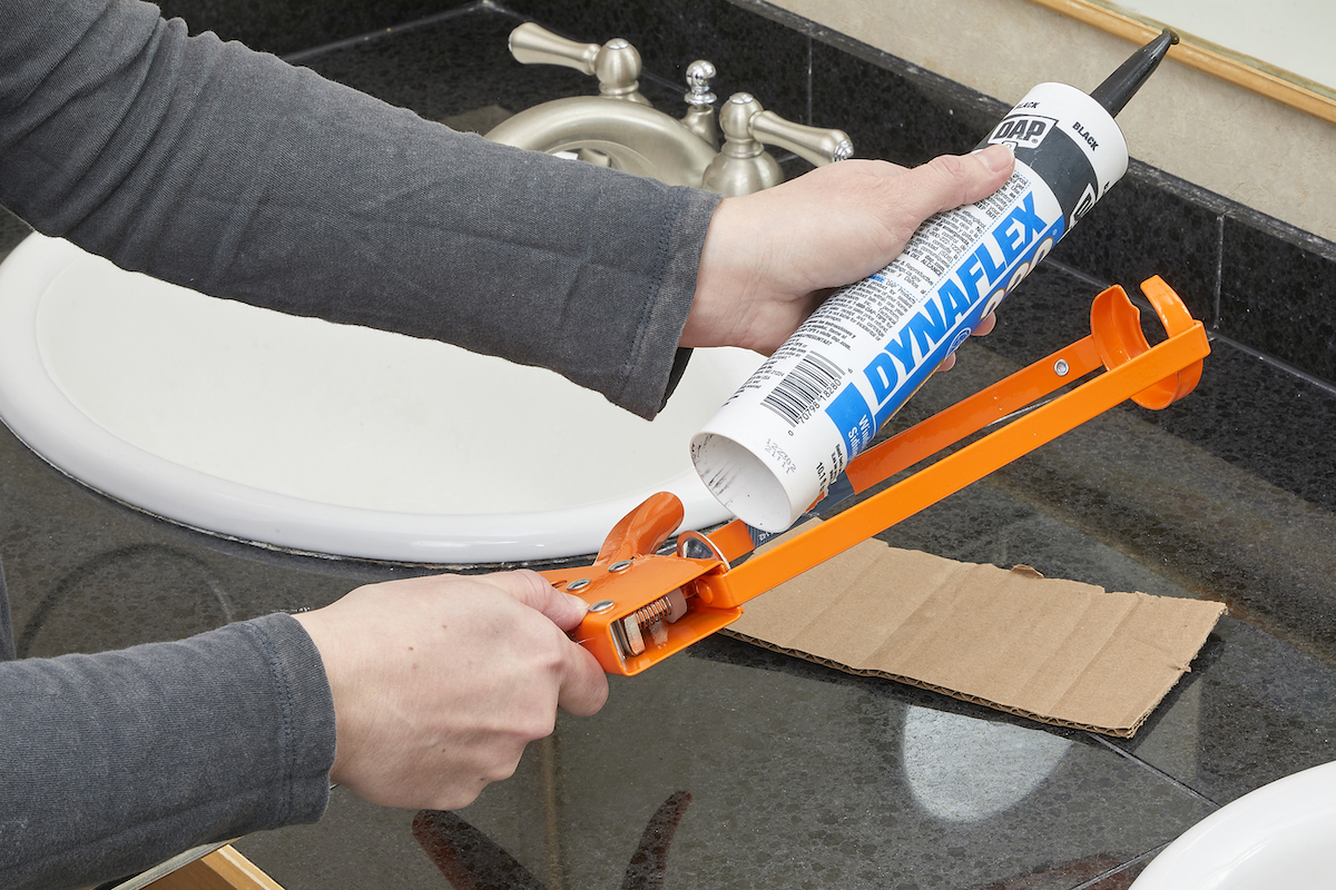 Woman inserts a tube of caulk into an orange caulk gun, with a bathroom vanity in the background.