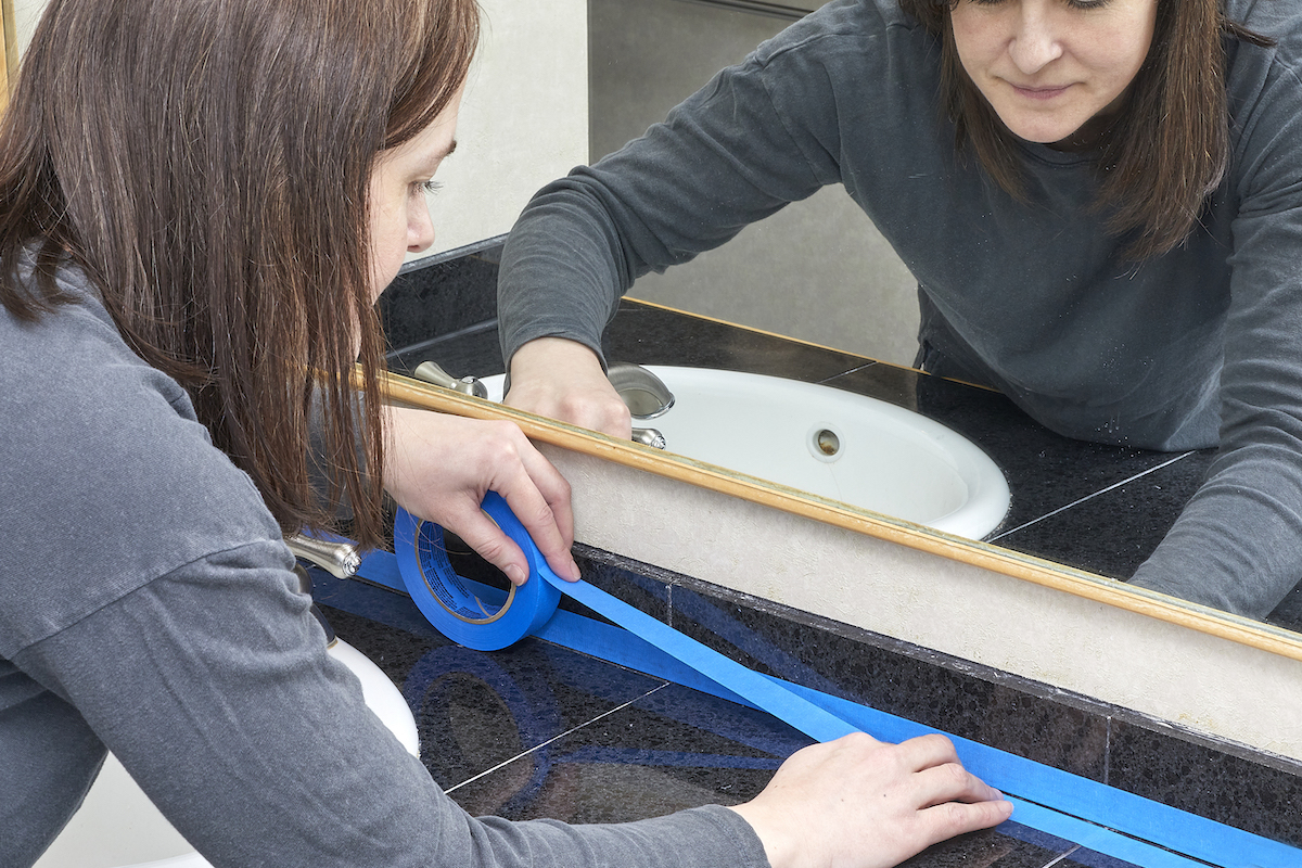 Woman tapes the caulking in front of her bathroom mirror with painter's tape.