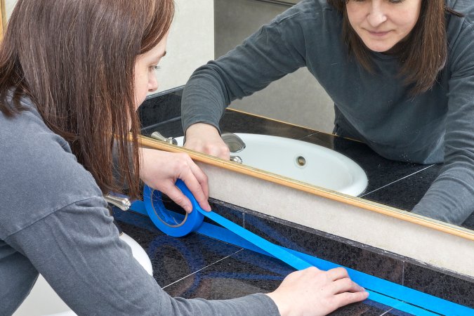 Woman tapes the caulking in front of her bathroom mirror with painter's tape.