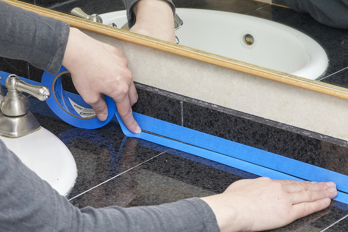 Woman applies painter's tape around caulk seams in front of her bathroom mirror.