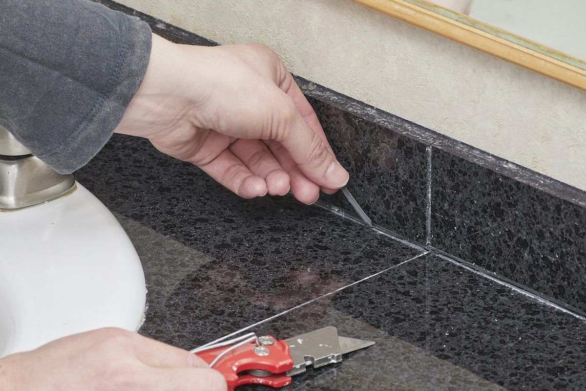 Woman pulls old caulk off of tile behind her bathroom sink; an X-acto knife is nearby.