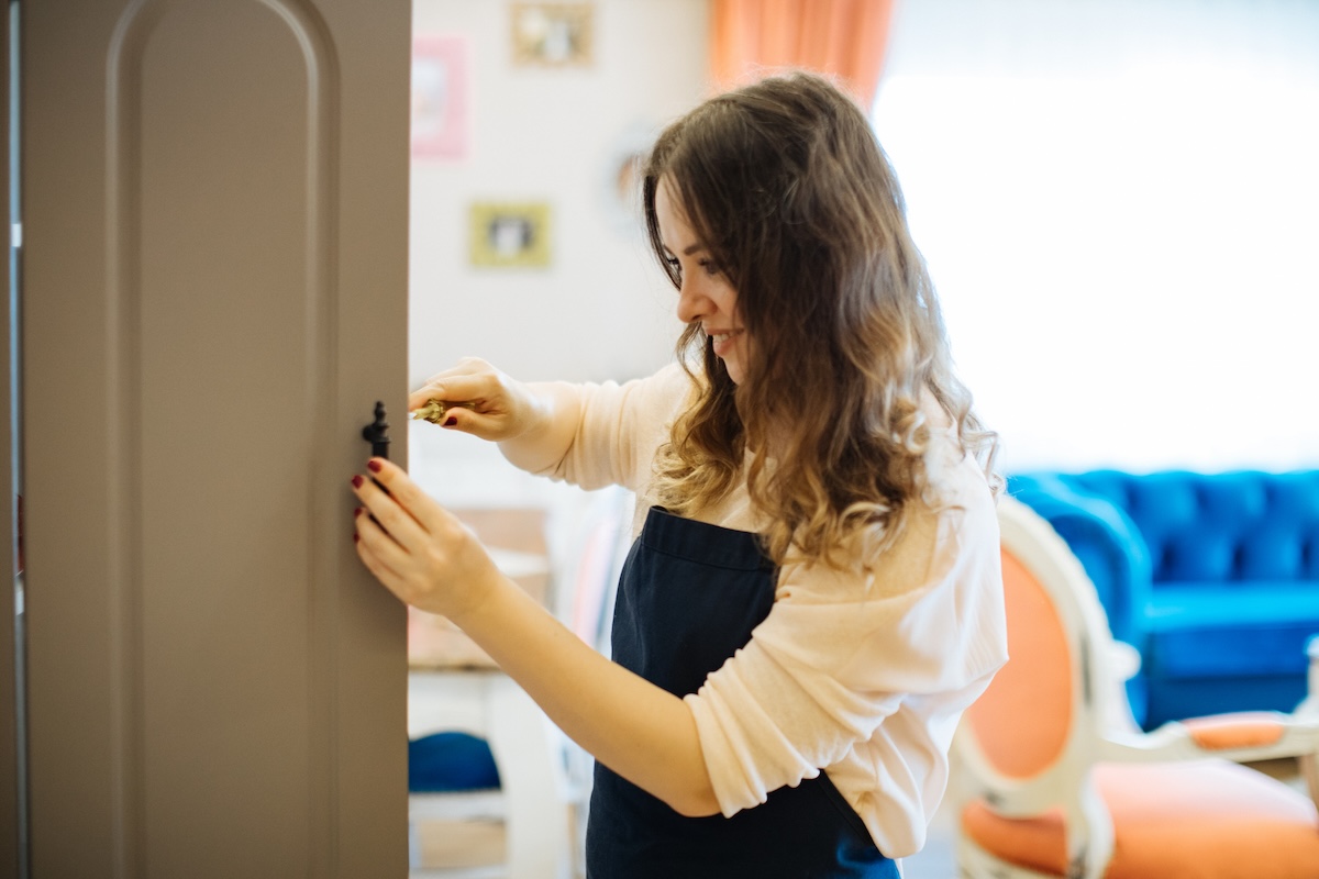 A young woman uses a screwdriver to remove handles from a painted cabinet. 