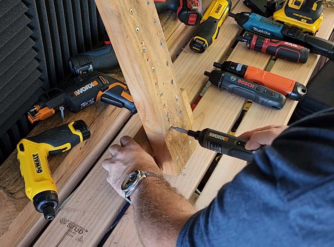 A person testing the best screwdriver option on a piece of wood with several other electric screwdrivers on a table in the background.