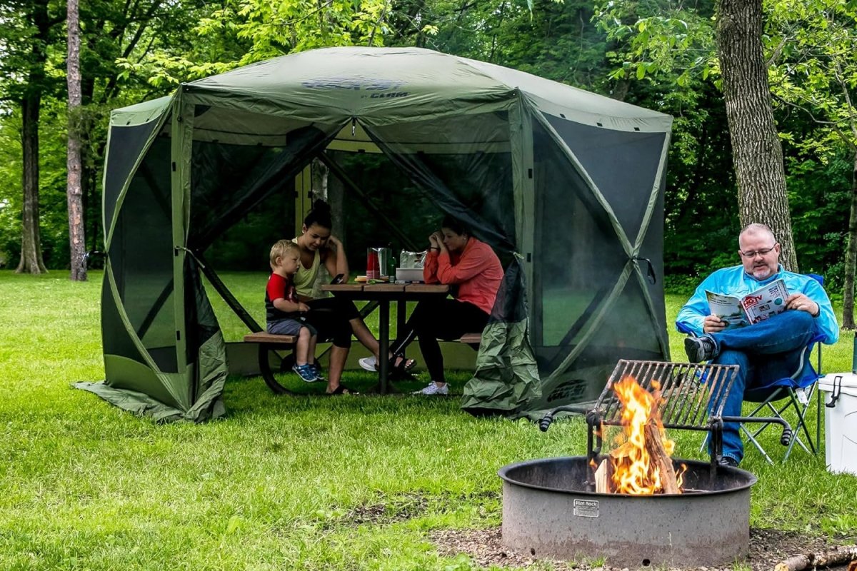 Two women and a child sitting under a canopy outside on the grass, next to a man sitting near a fire pit