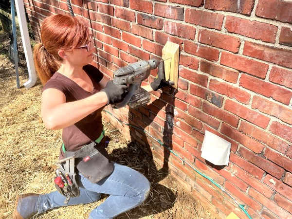 A woman using a drill to drill a piece of wood into a brick wall.