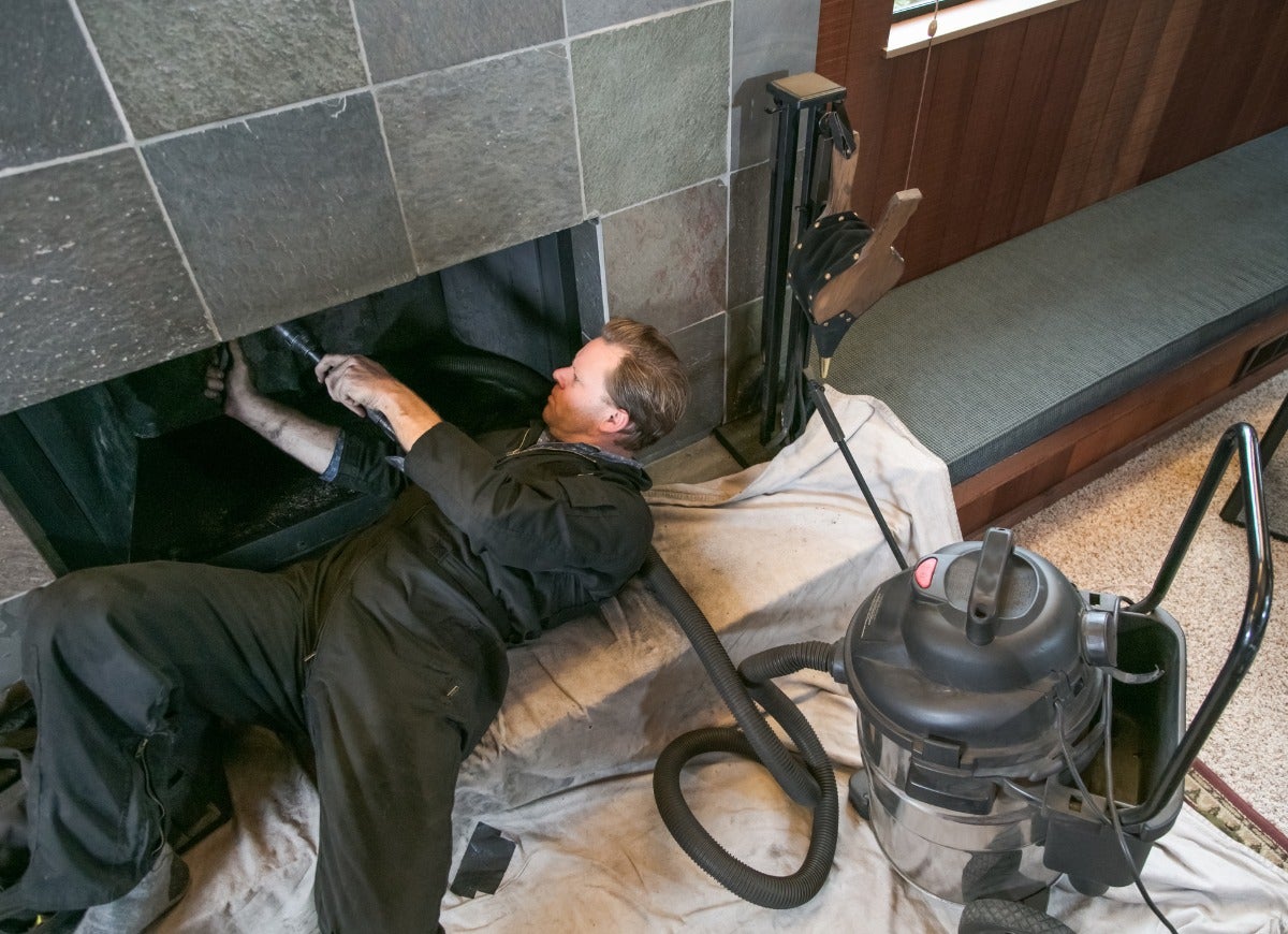 A repair person with a black jumpsuit lays on their back to inspect the inside of a fireplace.