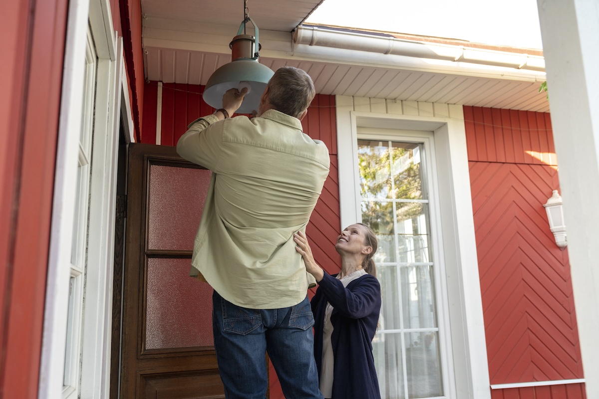 An older man on a ladder changes the lightbulb on a porch light attached to a red house.