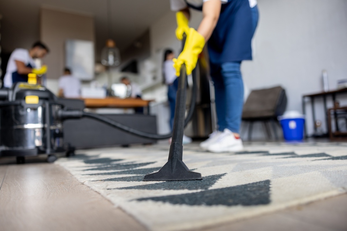 A professional wearing yellow gloves guides a steamed carpet cleaner across a white and gray rug.