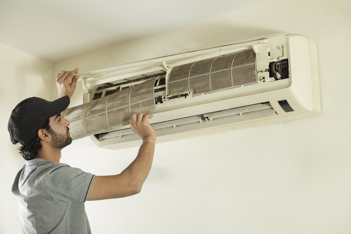 A man in a gray shirt and baseball cap repairs an indoor HVAC unit.