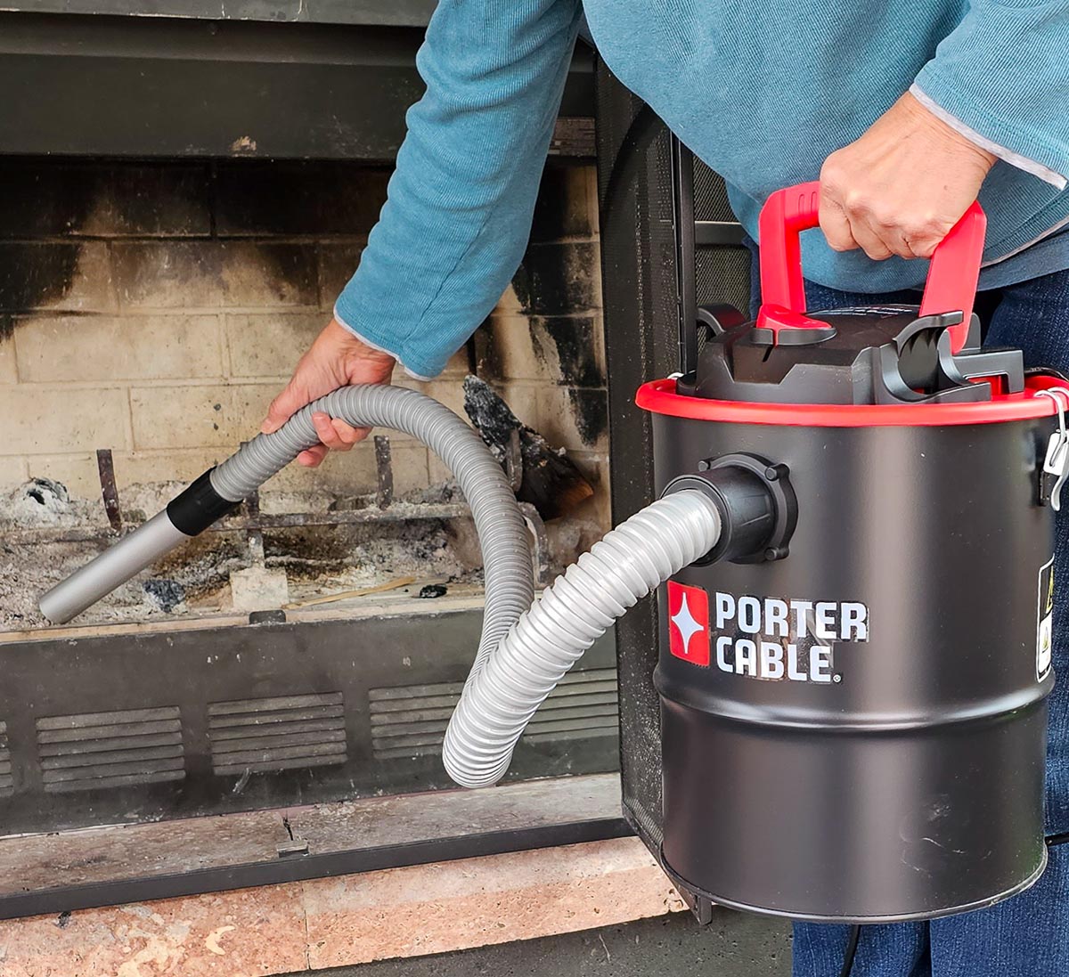 A person holding the best ash vacuum in one hand while vacuuming ash from an indoor fireplace with the other.