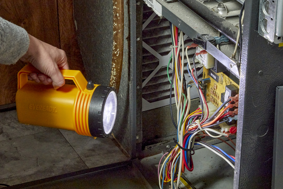 Woman shines a large flashlight into the mechanics of a furnace, whose door is open.