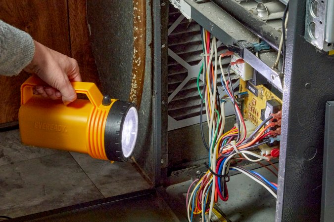 Woman shines a large flashlight into the mechanics of a furnace, whose door is open.