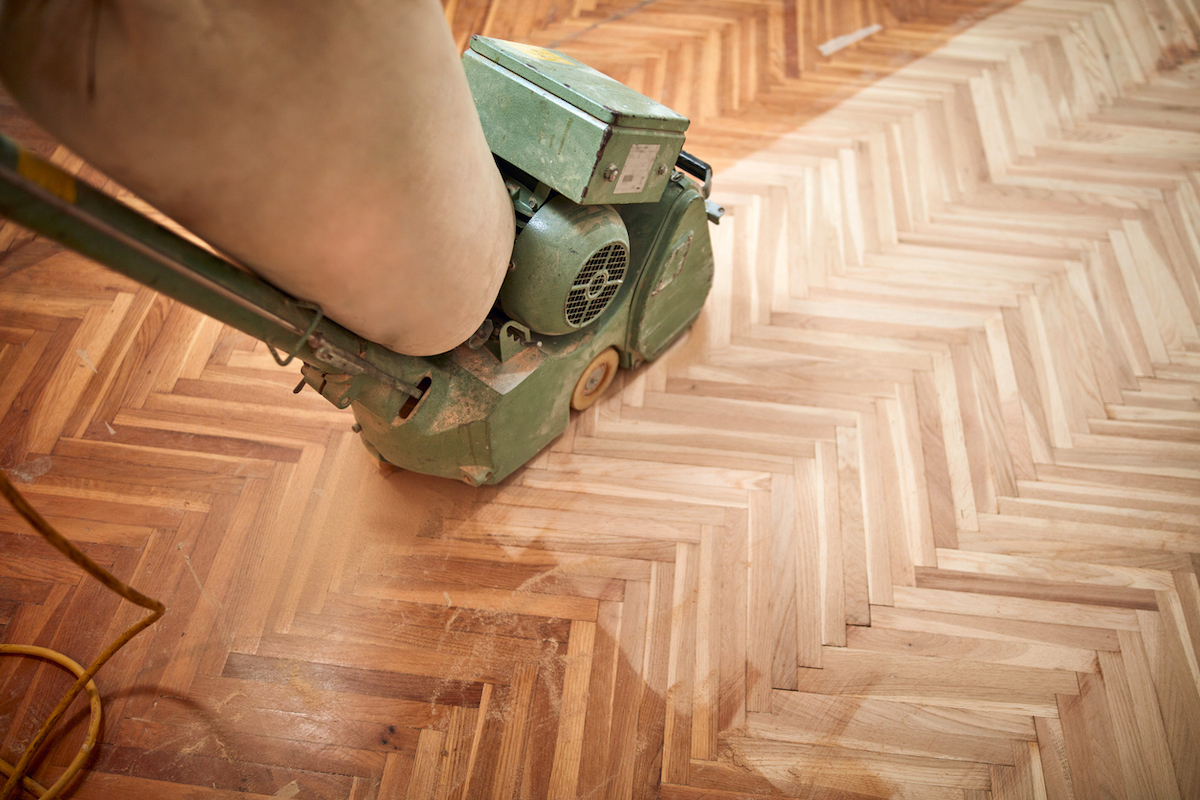 A floor sander is being used to sand hardwood flooring that has a herringbone pattern.