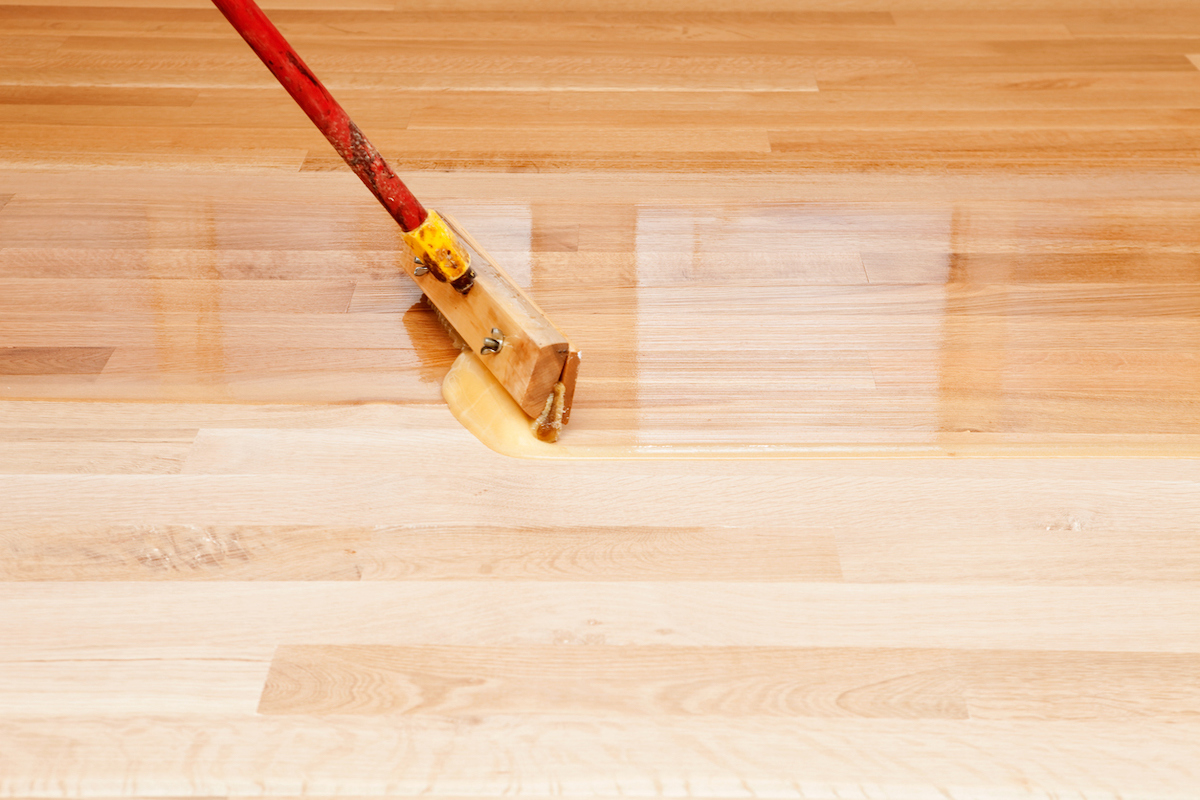 A wax applicator brush is being used to apply clear polyurethane to a sanded hardwood floor.