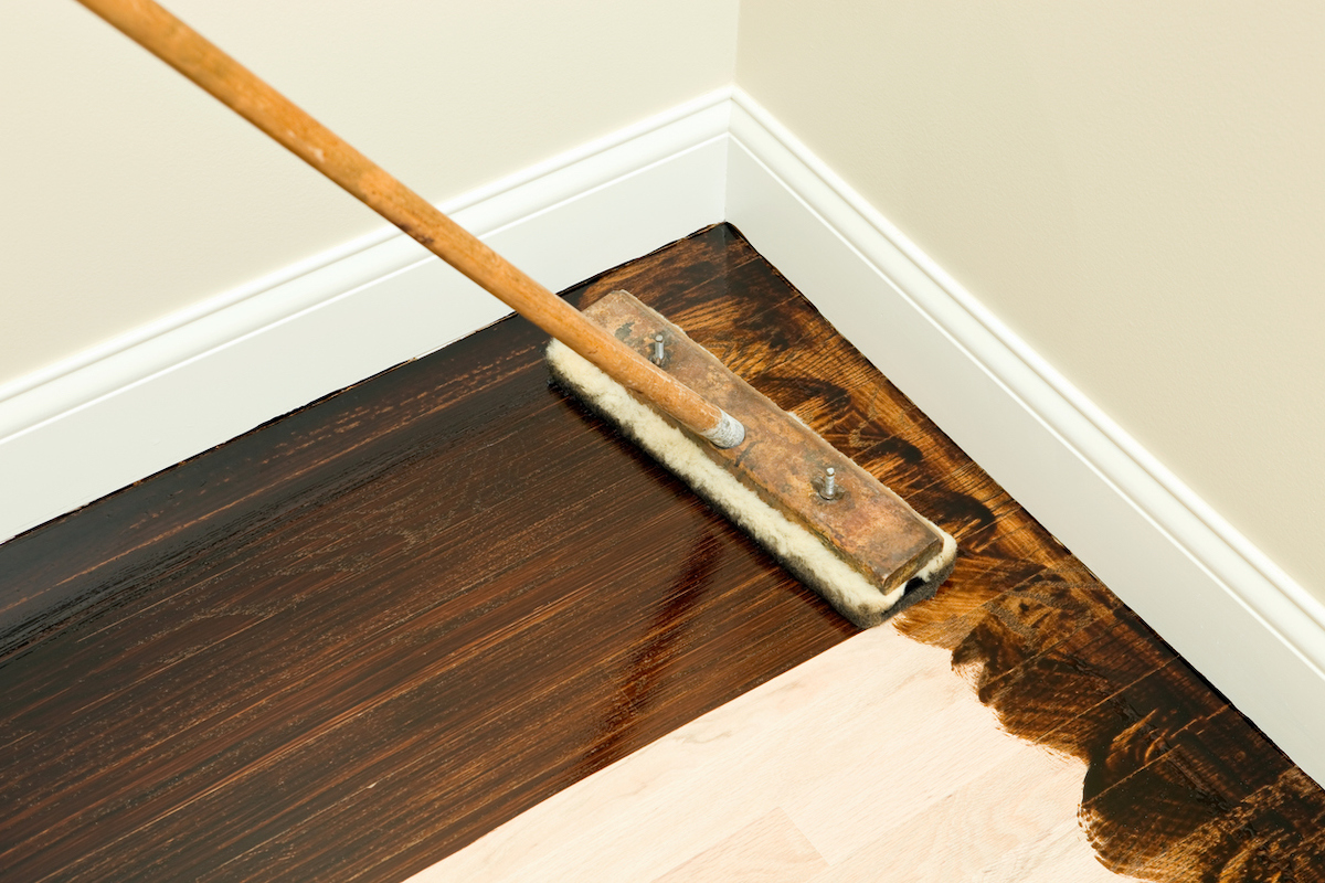 A long applicator brush is being used to apply a dark wood stain to sanded hardwood flooring.