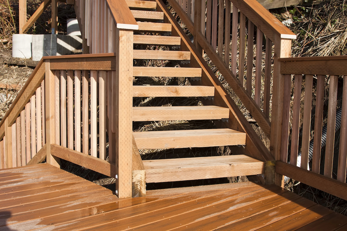 A wet wooden deck has railing with steps leading down to the ground and stairs leading up to another level unseen.