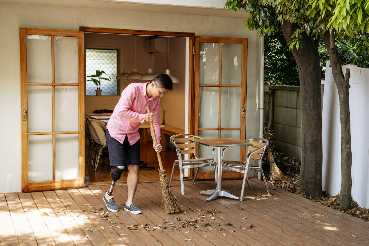 A person with a prosthetic leg is sweeping a wooden deck in a back yard.