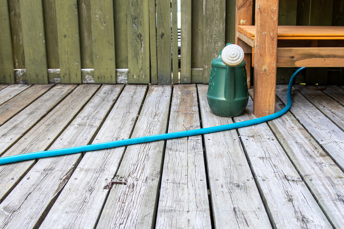 A wooden table, a green watering can, and a blue garden hose are on top of a wooden deck.