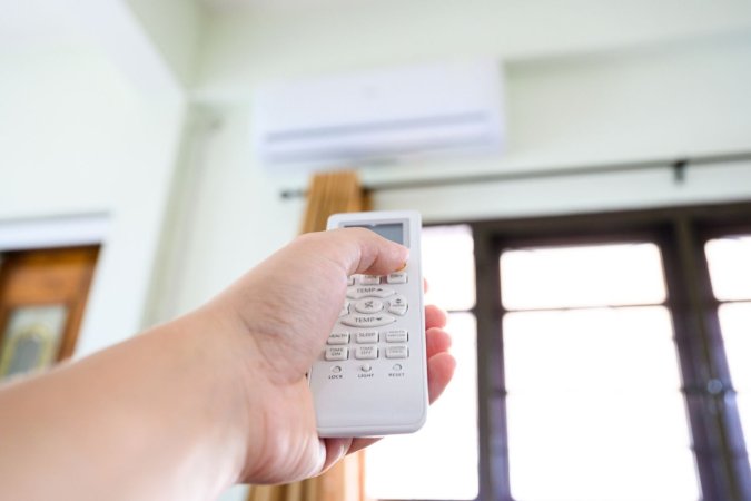 A person using a remote to adjust the best heat pump option mounted on a wall above a window