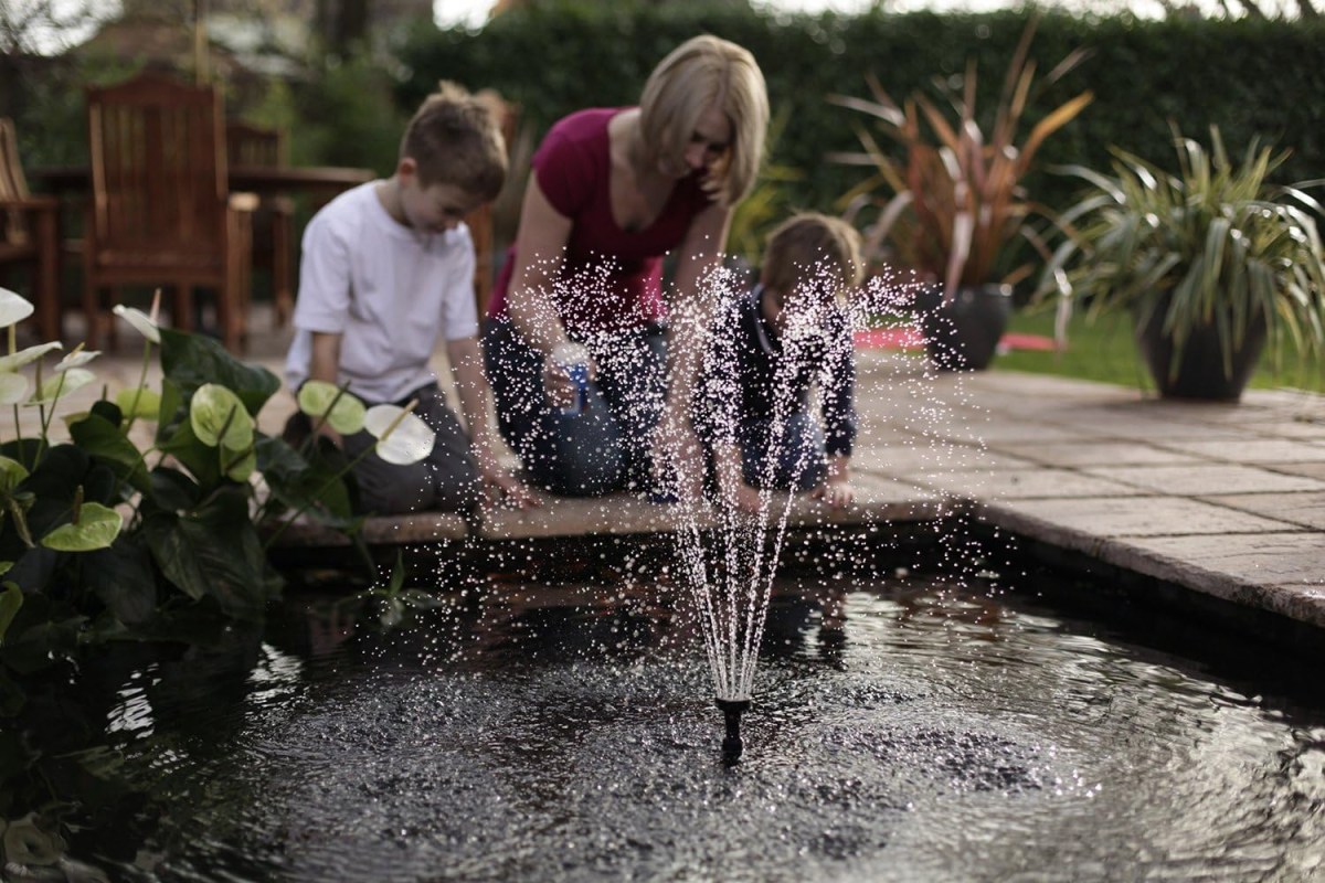A woman and 2 children kneeling next to a pond with a fountain spraying water