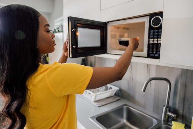 A young woman places container of food into black microwave in a kitchen.