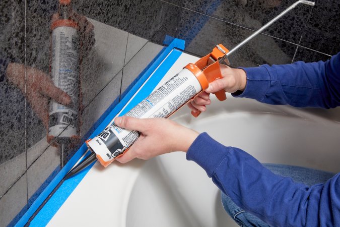 Person holding a caulk gun caulks an area around a bathtub marked with painter's tape.