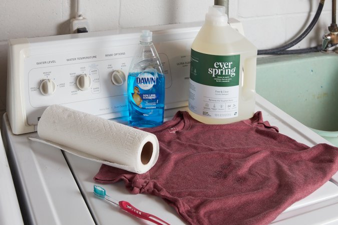 A rose-colored T-shirt and materials needed to clean it, including detergent, dish soap, and paper towels.