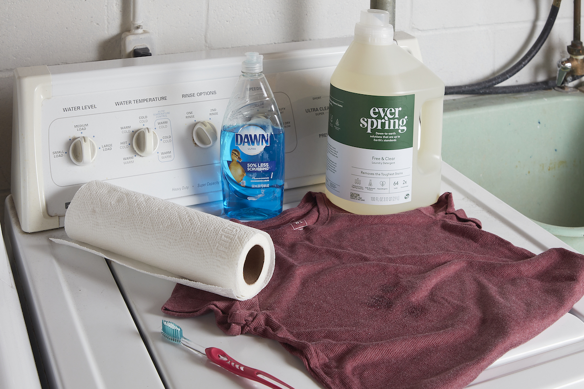 A rose-colored T-shirt and materials needed to clean it, including detergent, dish soap, and paper towels.