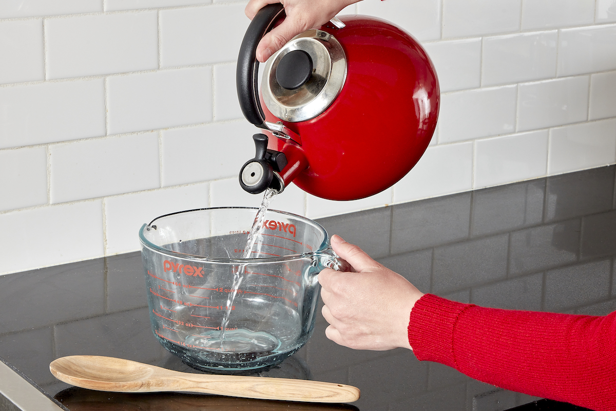 Woman pouring water from red teakettle into a glass measuring cup.