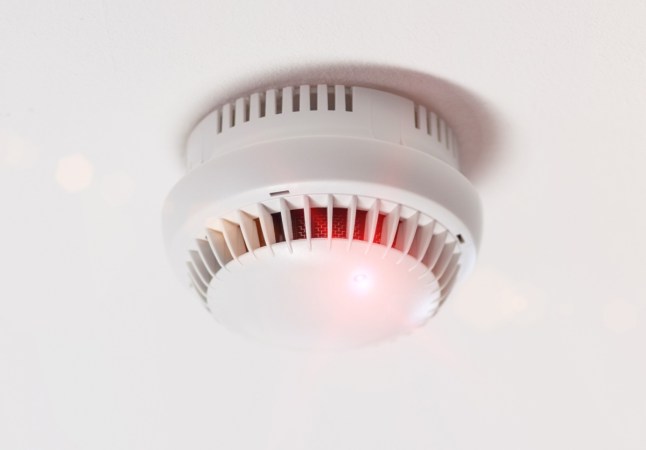 A smoke alarm installed on the ceiling has a red light on.
