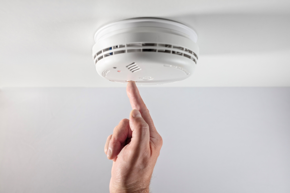 A man's hand is pressing the test button on the ceiling smoke alarm.