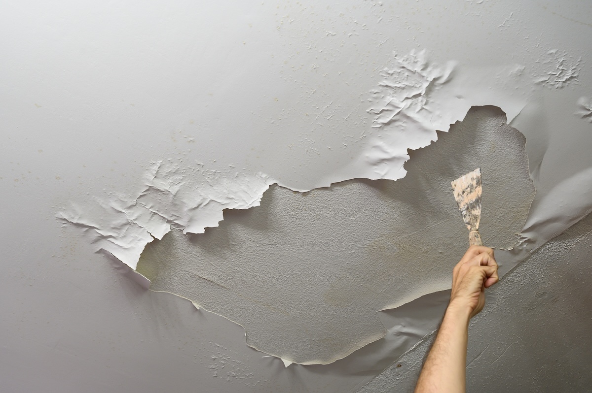 A man's hand scapes bubbled paint from a ceiling. 