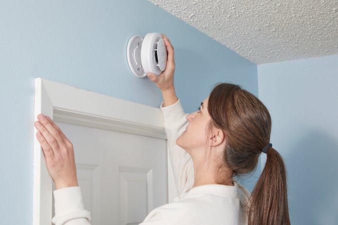 Woman pulls a smoke detector off its bracket, which is positioned above a doorframe.