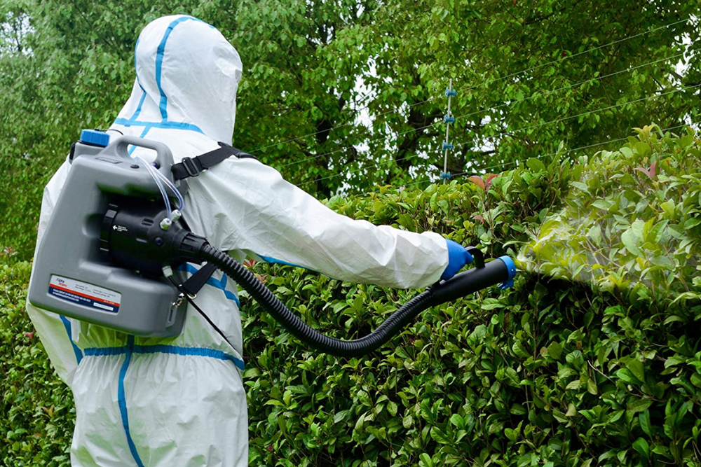 Man in coveralls using a backpack sprayer on a hedge