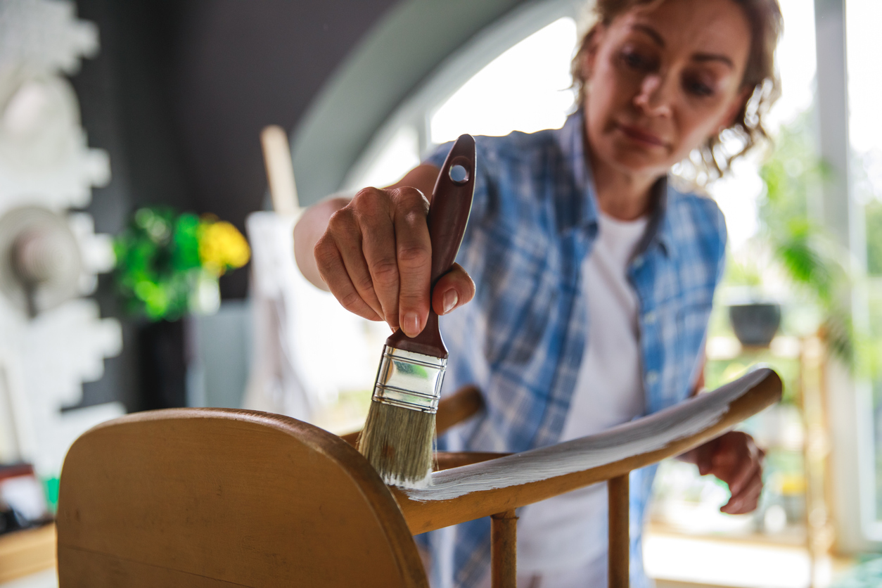 Focus on foreground of mature woman painting a chair with white paint while learning a new hobbie during lockdown days at home.