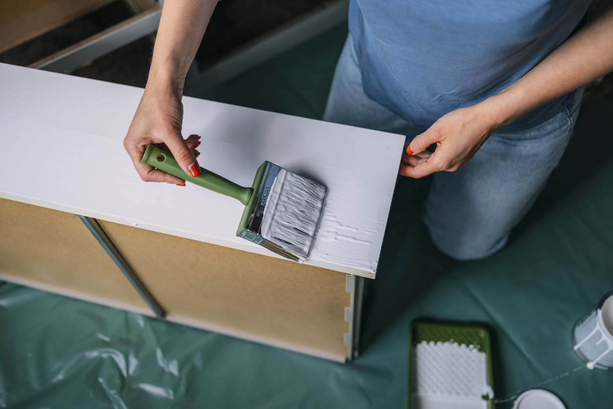 overhead view of hands and torso of woman with red fingernails applying a second coat of white paint to a dresser drawer