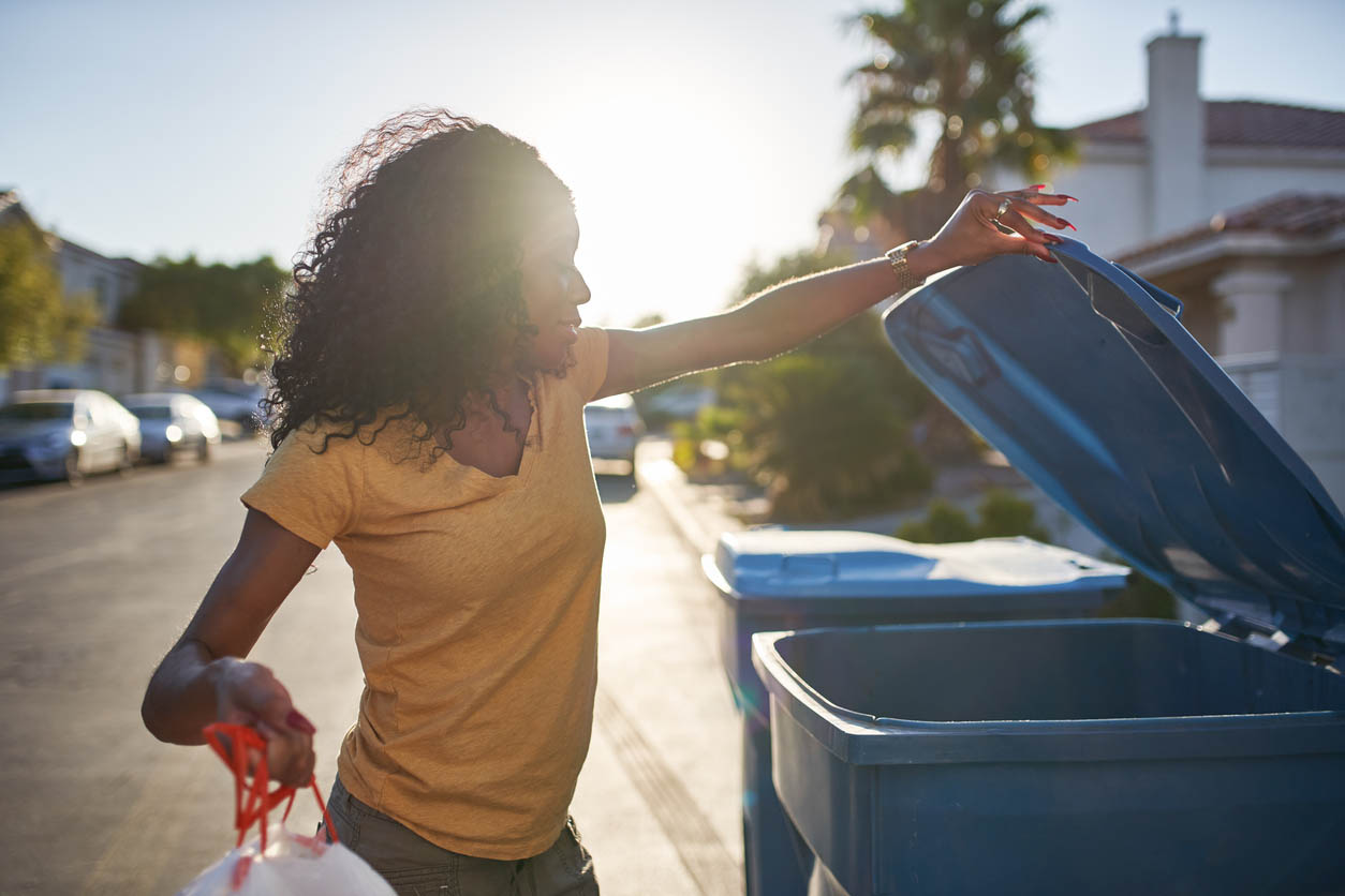 A person throws a bag of trash into an outdoor garbage can in a suburban neighborhood.