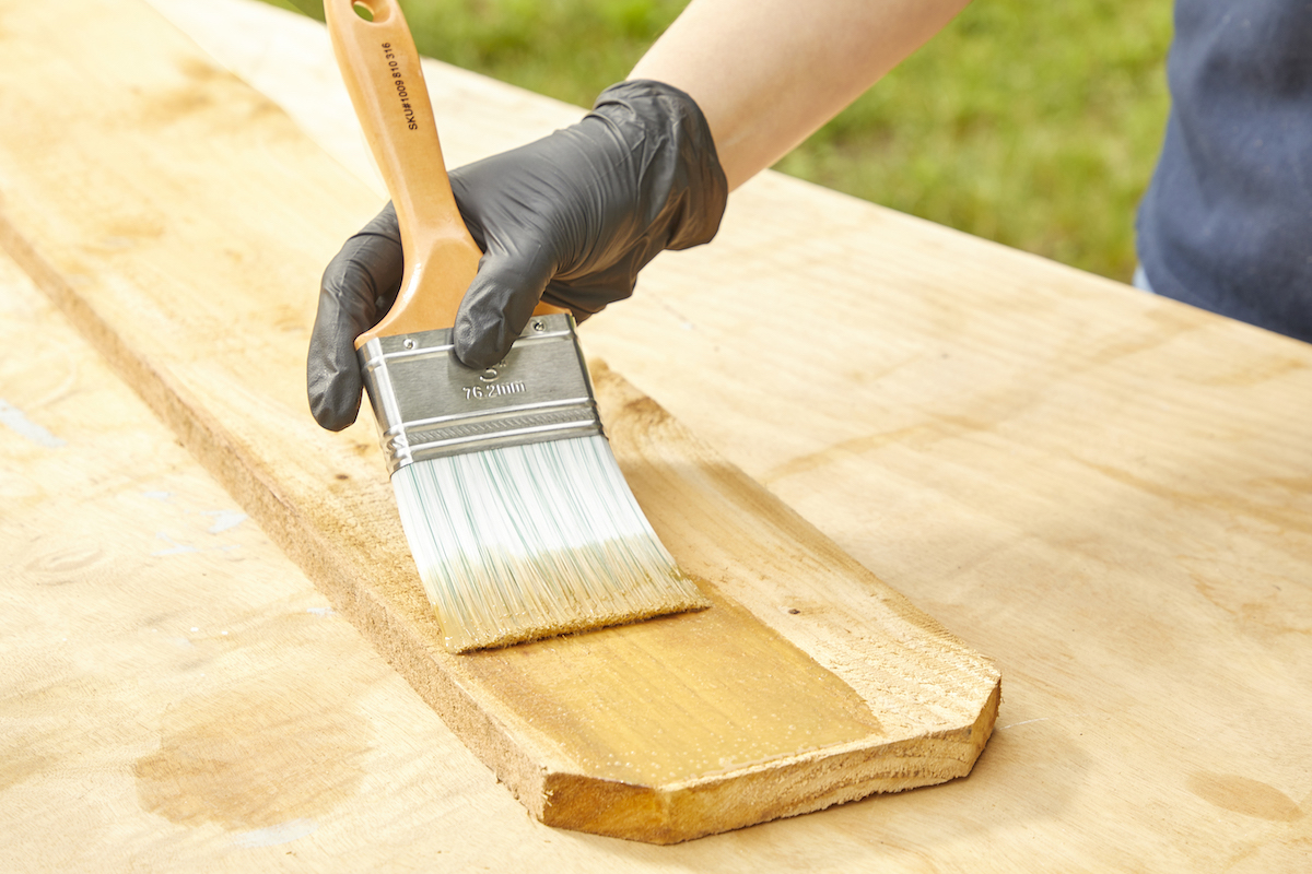 Woman uses paint brush to stain a board of pressure-treated wood.