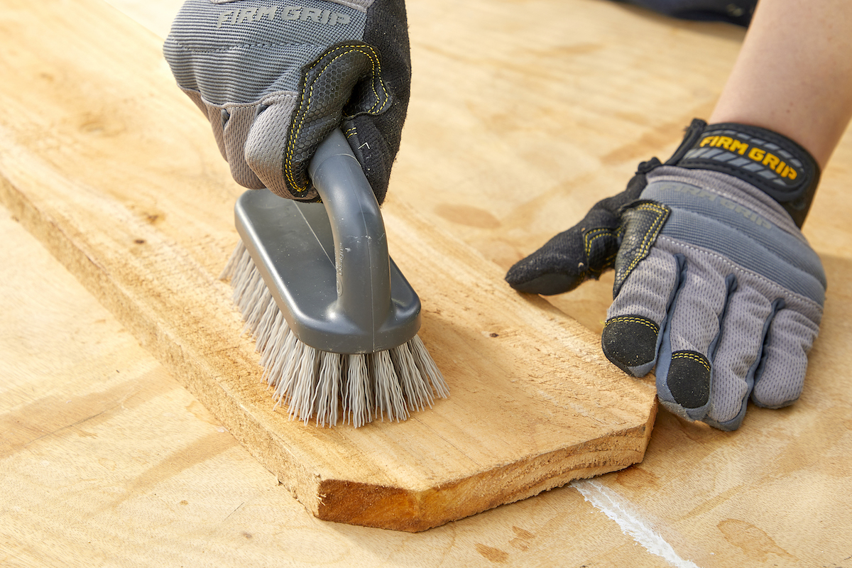 Woman wearing work gloves uses a scrub brush to clean a board of pressure-treated wood.