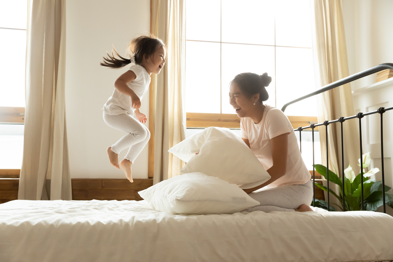 In morning carefree small daughter jump on bed while vietnamese mother laughing feels happy, asian ethnicity family in comfortable pyjamas wake up start new day positive mood enjoy active life concept