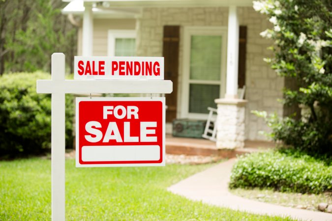 Red and white "Sale Pending, Home for Sale" sign in front of a stone, wood house that is for sale and has recently been sold. Green grass and bushes indicate the spring or summer season. Front porch and windows in background. Real estate signs in residential neighborhood. Moving house, relocation concepts.