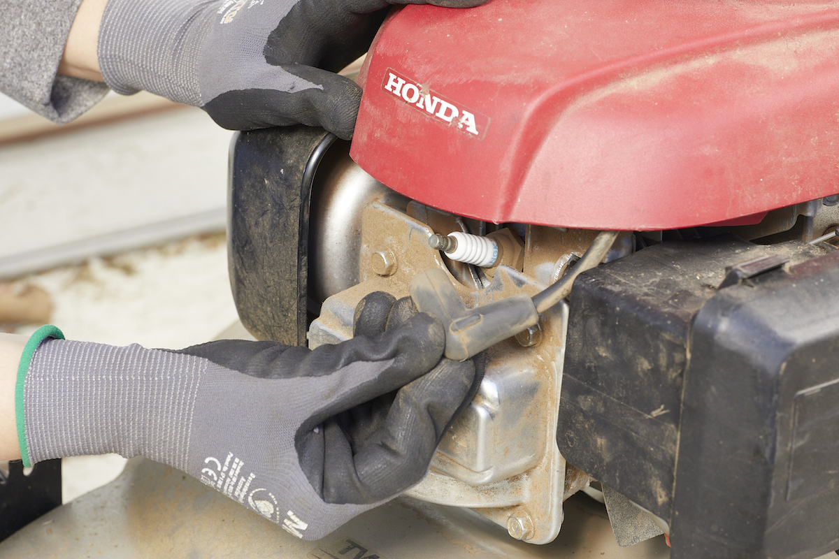 Person wearing work gloves exposes spark plug on lawn mower.